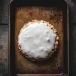 Oatmeal cookie glaze - the image shows a square baking tray with a dessert on it. the dessert appears to be a round, golden-brown crust with a crumbly texture. on top of the crust, there is a layer of white icing that is drizzled over the top. the icing is smooth and shiny, and it looks like it has been freshly baked. the tray is placed on a dark wooden surface, and the background is blurred, making the dessert the focal point of the image.