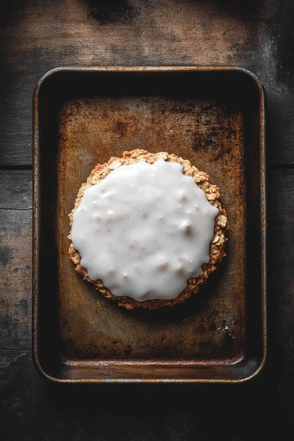 Oatmeal cookie glaze - the image shows a square baking tray with a dessert on it. the dessert appears to be a round, golden-brown crust with a crumbly texture. on top of the crust, there is a layer of white icing that is drizzled over the top. the icing is smooth and shiny, and it looks like it has been freshly baked. the tray is placed on a dark wooden surface, and the background is blurred, making the dessert the focal point of the image.