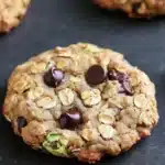Oatmeal zucchini chocolate chip cookie - the image shows a close-up of a freshly baked oatmeal cookie on a black baking tray. the cookie is round and golden brown in color, with chunks of chocolate and nuts scattered throughout. it appears to be soft and chewy, with a slightly crumbly texture. in the background, there are more cookies, but they are slightly blurred. the tray is lined with parchment paper, and there is a metal spatula resting on the right side of the image.