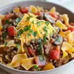 One pot beef noodle skillet - the image is a close-up of a dish in a gray bowl. the dish appears to be a type of pasta dish, possibly fettuccine alfredo, with a layer of ground beef, diced tomatoes, mushrooms, and cheese on top. the pasta is cooked al dente and is arranged in a circular pattern around the bowl. there is a sprig of fresh parsley on top of the dish, adding a pop of green color to the dish. the bowl is sitting on a wooden table, and there are a few sprigs of parsley scattered around the table.