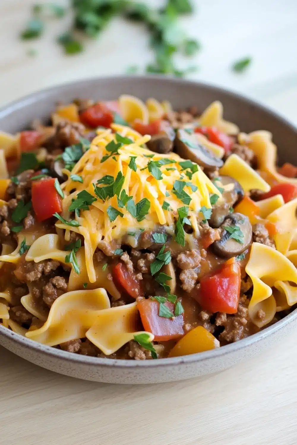 One pot beef noodle skillet - the image is a close-up of a dish in a gray bowl. the dish appears to be a type of pasta dish, possibly fettuccine alfredo, with a layer of ground beef, diced tomatoes, mushrooms, and cheese on top. the pasta is cooked al dente and is arranged in a circular pattern around the bowl. there is a sprig of fresh parsley on top of the dish, adding a pop of green color to the dish. the bowl is sitting on a wooden table, and there are a few sprigs of parsley scattered around the table.