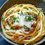 One pot carmelized onion pasta - the image is a close-up of a bowl of spaghetti. the spaghetti is cooked al dente and is topped with a generous amount of grated parmesan cheese. the dish is garnished with fresh parsley leaves. the bowl is made of dark brown ceramic and is sitting on a dark grey surface. the background is blurred, but it appears to be a kitchen countertop.