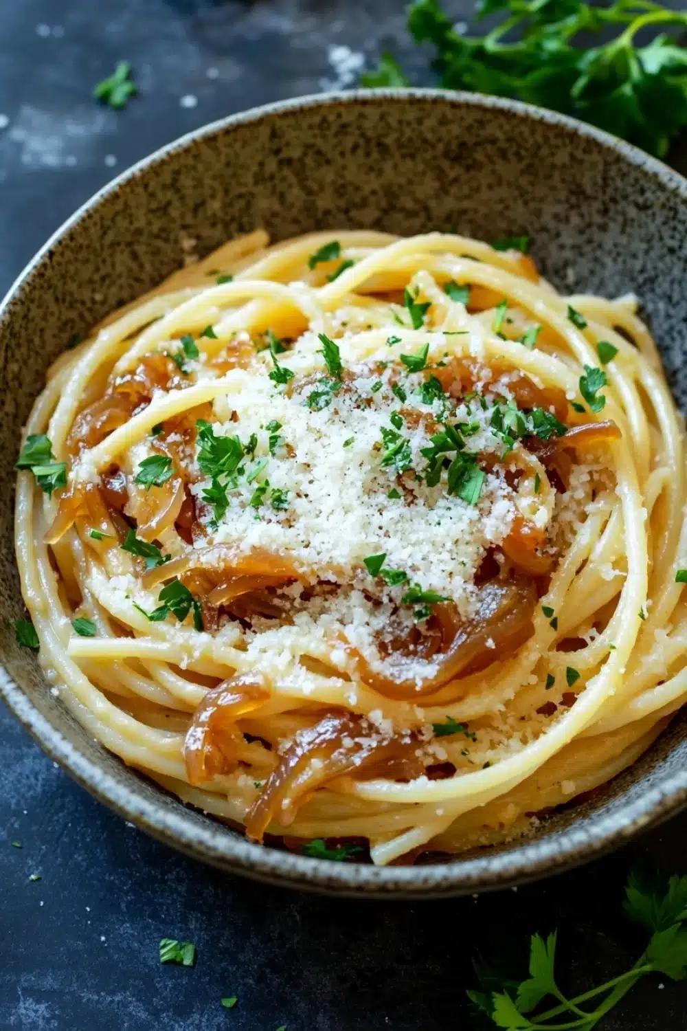 One pot carmelized onion pasta - the image is a close-up of a bowl of spaghetti. the spaghetti is cooked al dente and is topped with a generous amount of grated parmesan cheese. the dish is garnished with fresh parsley leaves. the bowl is made of dark brown ceramic and is sitting on a dark grey surface. the background is blurred, but it appears to be a kitchen countertop.