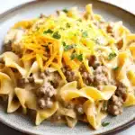 One pot creamy beef and noodles - the image shows a plate of pasta dish with a creamy sauce and chunks of ground beef. the pasta appears to be fettuccine alfredo, which is a type of spiral-shaped pasta. the sauce is a light brown color and is drizzled over the top of the dish. the dish is garnished with chopped parsley and a sprinkle of grated cheese. the plate is sitting on a white marble countertop.