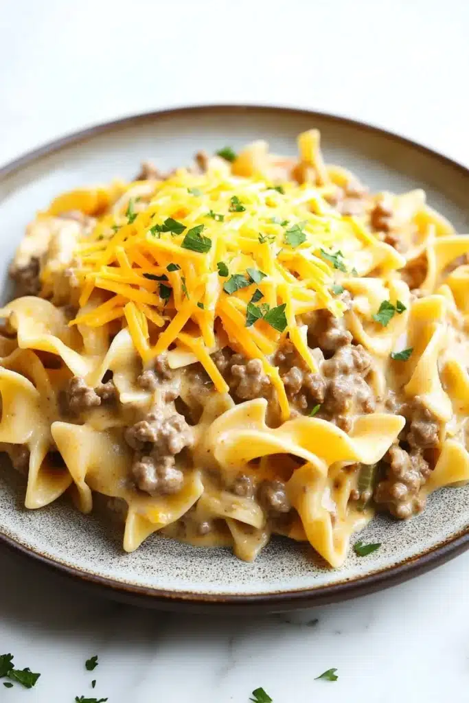One pot creamy beef and noodles - the image shows a plate of pasta dish with a creamy sauce and chunks of ground beef. the pasta appears to be fettuccine alfredo, which is a type of spiral-shaped pasta. the sauce is a light brown color and is drizzled over the top of the dish. the dish is garnished with chopped parsley and a sprinkle of grated cheese. the plate is sitting on a white marble countertop.