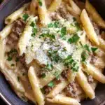 One pot creamy garlic butter beef pasta - the image shows a bowl of penne pasta dish. the pasta is cooked al dente and is topped with a creamy sauce and chunks of ground meat. the sauce is a light beige color and appears to be thick and creamy. the dish is garnished with chopped parsley and grated parmesan cheese. the bowl is made of dark brown ceramic and is sitting on a wooden table. there are a few sprigs of parsley scattered around the bowl.