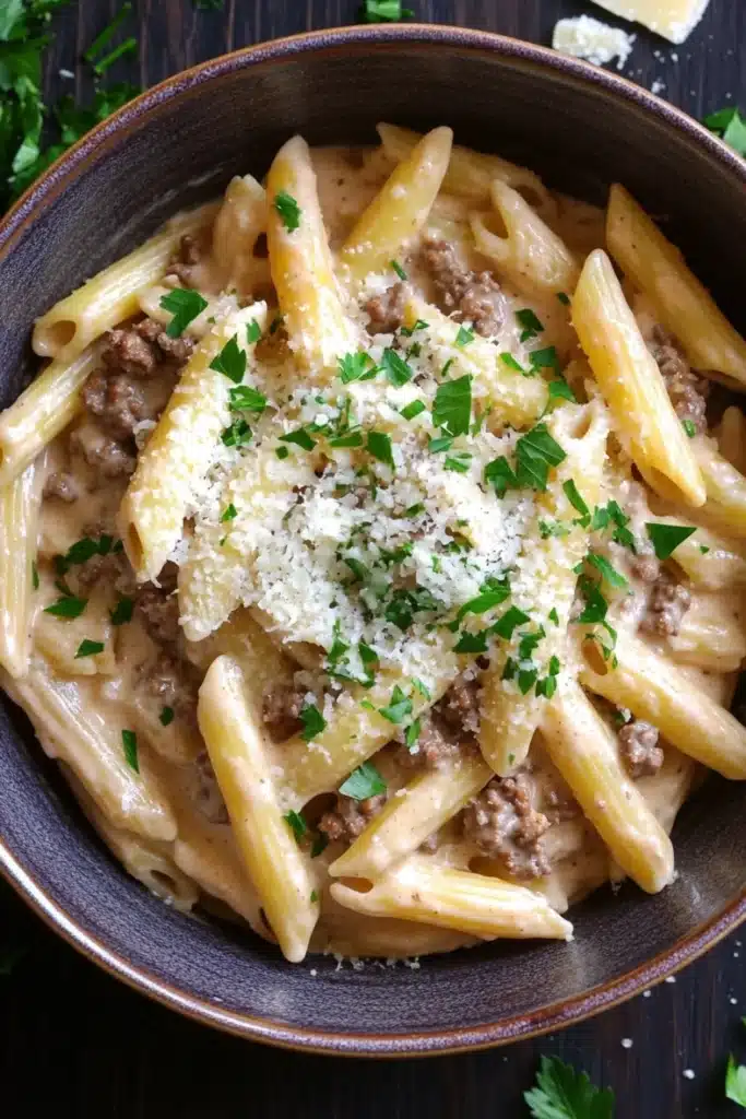 One pot creamy garlic butter beef pasta - the image shows a bowl of penne pasta dish. the pasta is cooked al dente and is topped with a creamy sauce and chunks of ground meat. the sauce is a light beige color and appears to be thick and creamy. the dish is garnished with chopped parsley and grated parmesan cheese. the bowl is made of dark brown ceramic and is sitting on a wooden table. there are a few sprigs of parsley scattered around the bowl.