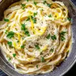 One pot creamy lemon pasta - the image is a close-up of a bowl of tagliatelle pasta. the pasta is cooked al dente and is topped with grated parmesan cheese and fresh parsley. the bowl is made of ceramic and has a dark brown rim. the background is a dark grey textured surface. the overall color scheme of the image is warm and inviting.