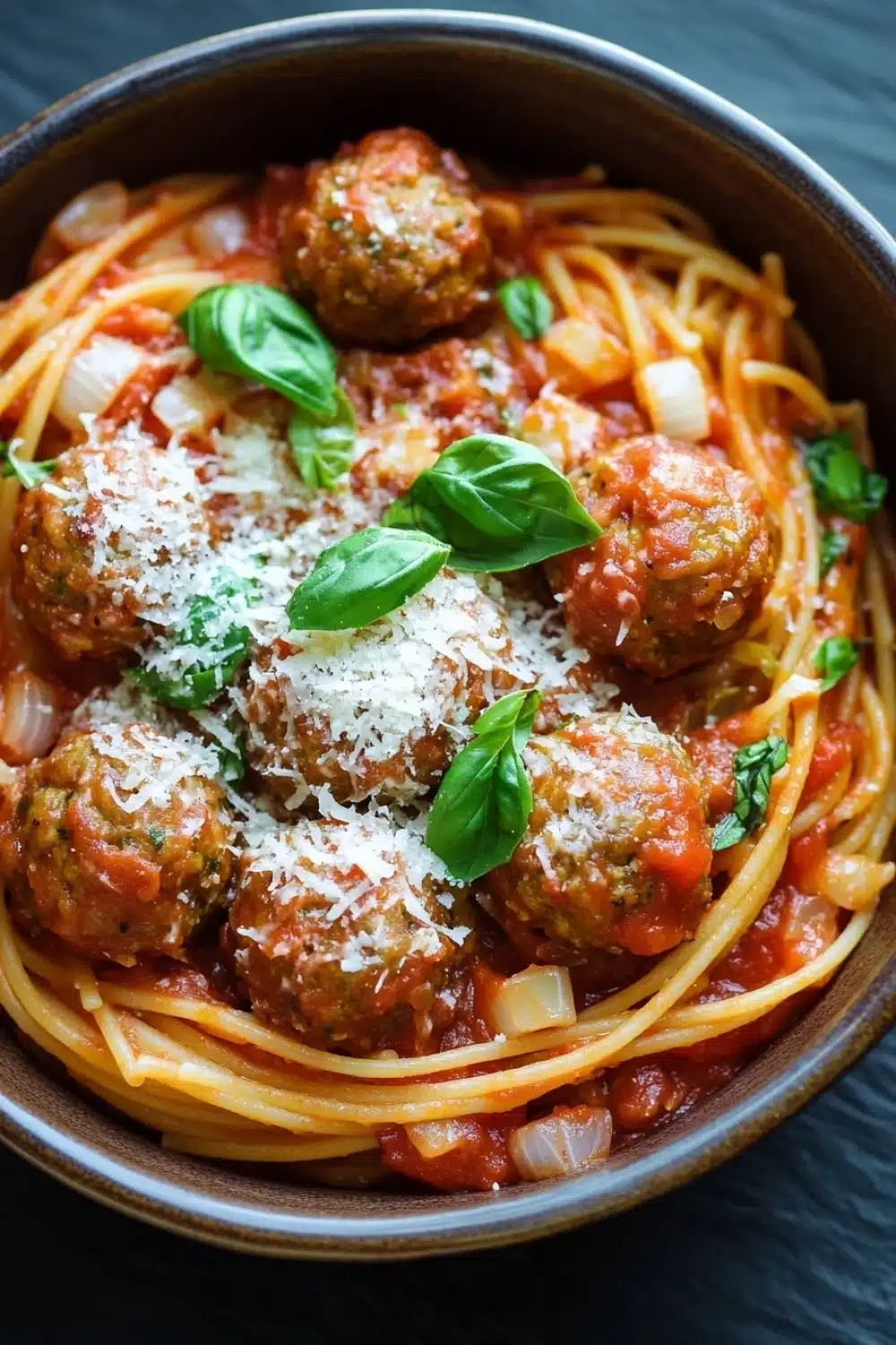 One pot pasta and meatballs - the image is a close-up of a bowl of spaghetti and meatballs. the spaghetti is cooked al dente and is covered in a tomato-based sauce with chunks of meat and vegetables. the meatballs are golden brown and appear to be seasoned with grated parmesan cheese. there are also some fresh basil leaves scattered on top of the dish. the bowl is made of ceramic and is sitting on a dark blue tablecloth.