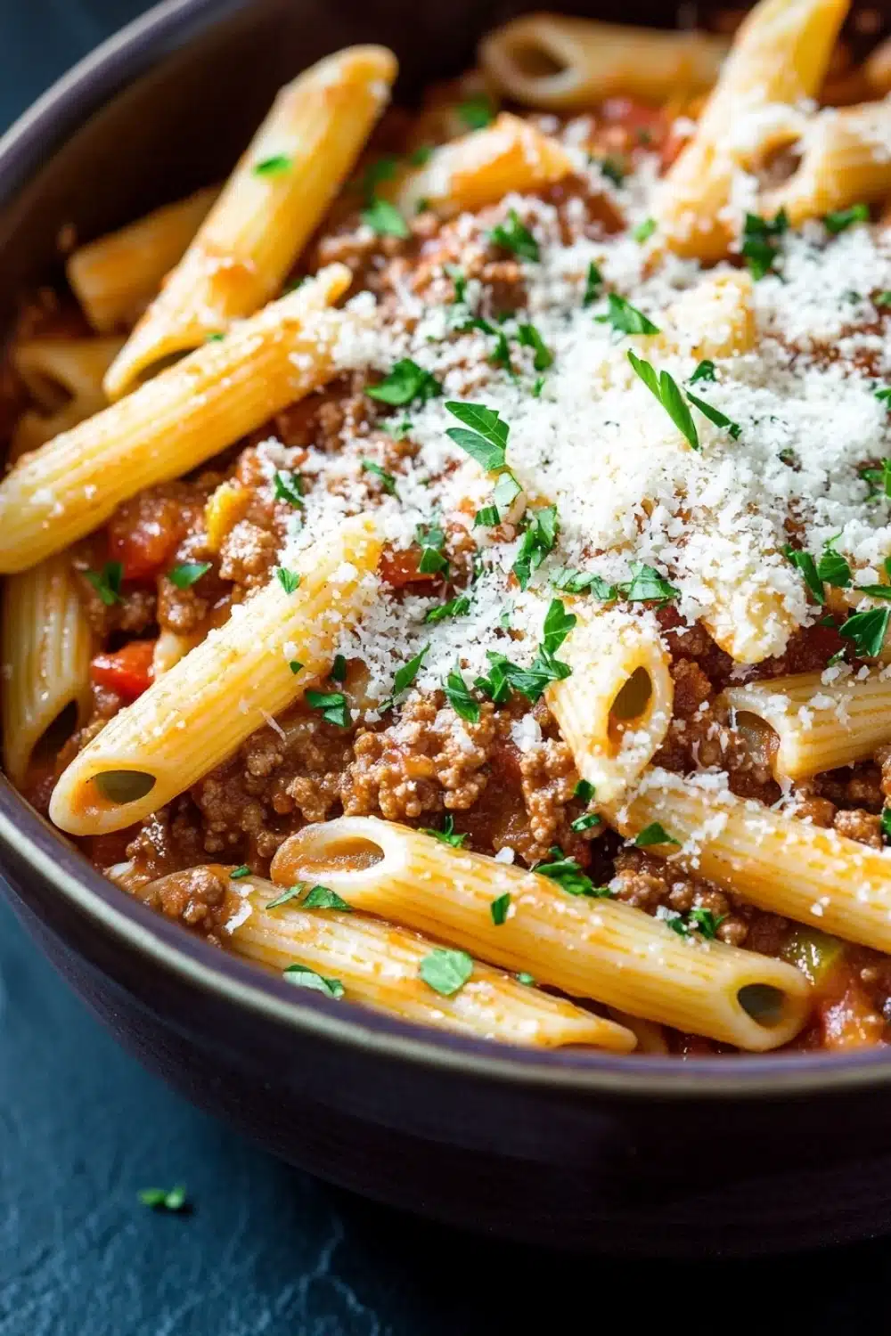 One pot pasta with meat sauce - the image shows a close-up of a bowl of pasta dish. the pasta appears to be penne, which is a type of penne pasta, and it is cooked in a tomato-based sauce with chunks of ground meat and tomato sauce. the dish is garnished with chopped parsley and grated parmesan cheese. the bowl is made of dark brown ceramic and is sitting on a dark blue surface. the background is blurred, making the pasta the focal point of the image.