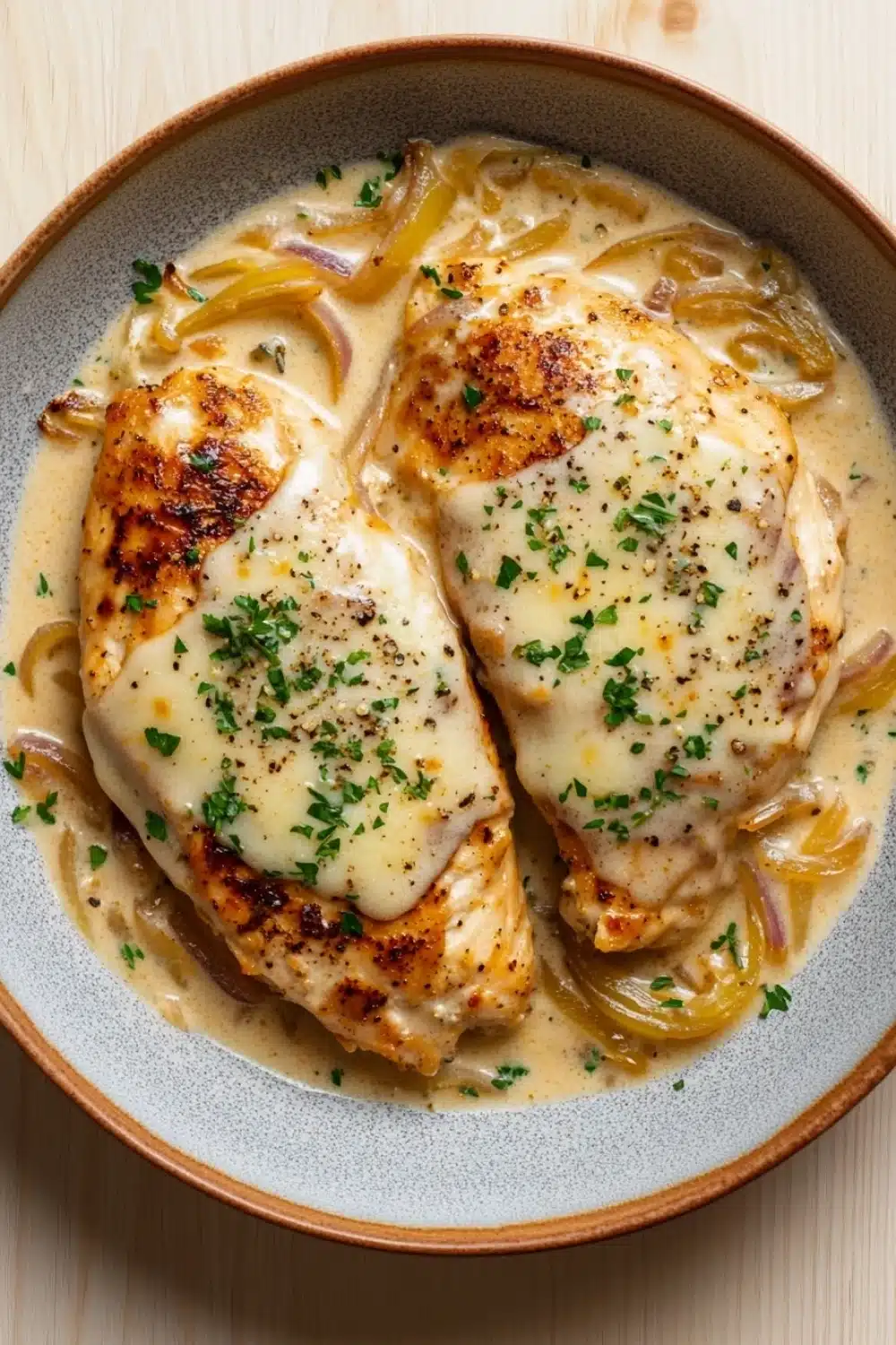 One skillet creamy french onion chicken - the image shows a plate of food with two pieces of chicken breasts covered in a creamy sauce. the chicken breasts are golden brown and appear to be seasoned with herbs and spices. the sauce is a light yellow color and is drizzled over the top of the chicken breasts. there are also chunks of caramelized onions scattered throughout the dish. the plate is made of ceramic and is sitting on a wooden table.