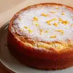 Orange french yogurt cake - the image is of a round cake on a blue plate. the cake appears to be freshly baked and has a golden brown crust. it is dusted with powdered sugar, giving it a shiny appearance. on top of the cake, there are small orange zest scattered around, adding a pop of color to the dish. the plate is sitting on a wooden table.
