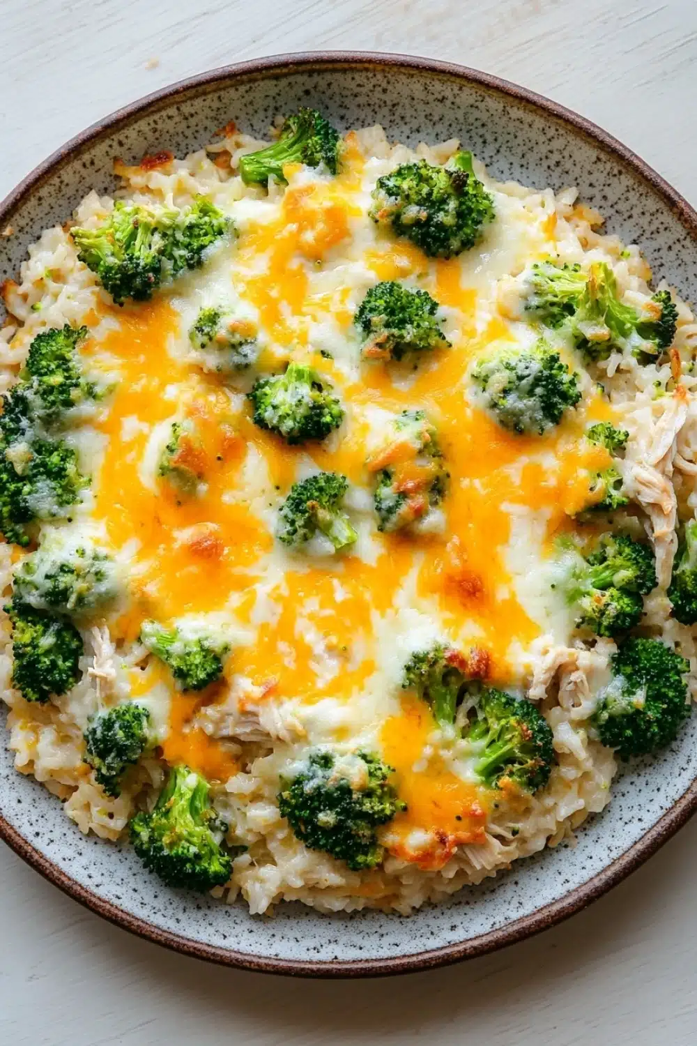 Oven baked chicken broccoli rice casserole - the image is a close-up of a dish in a bowl. the dish appears to be a casserole or risotto, with a layer of white rice and chunks of broccoli on top. the broccoli is cut into small pieces and is arranged in a circular pattern on top of the rice. the cheese is melted and bubbly, and it looks like it has been cooked until it is golden brown. the bowl is made of ceramic and has a brown rim. the background is a white wooden table.