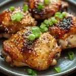Oven baked korean chicken thighs - the image shows a plate of grilled chicken breasts covered in sesame seeds and green onions. the chicken breasts are golden brown and appear to be seasoned with herbs and spices. the plate is round and has a dark grey rim. the background is blurred, but it appears to be a wooden table or countertop. the overall color palette of the image is warm and inviting.