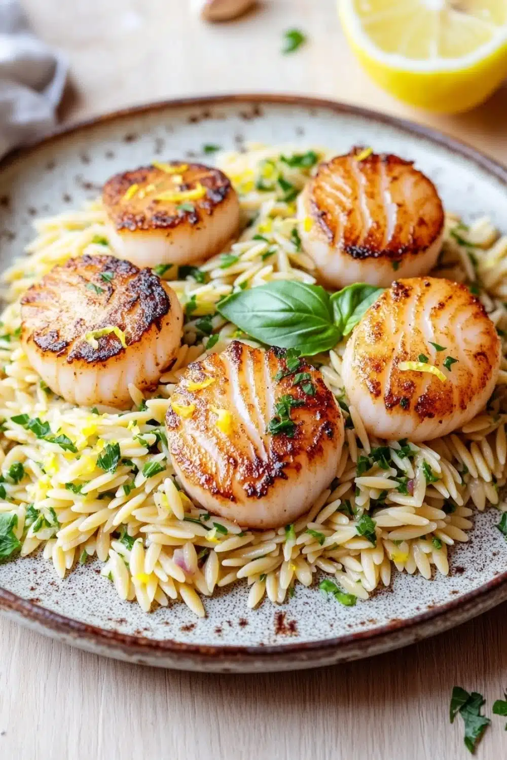 Pan seared scallops with orzo - the image shows a plate of grilled scallops on top of a bed of fusilli pasta. the scallop shells are golden brown and appear to be seasoned with herbs and spices. the pasta is a mix of yellow and white, and there are small pieces of lemon scattered throughout the dish. the plate is on a wooden table with a white cloth napkin and a lemon wedge in the background. the overall presentation of the dish is elegant and appetizing.