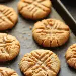 Peanut butter cookie no butter - the image shows a baking tray with freshly baked peanut butter cookies on it. the cookies are round and golden brown in color, with a criss-cross pattern on top. they are arranged in a single layer on the tray, with some overlapping each other. the tray is lined with parchment paper, and there is a small red bowl on the right side of the tray. the background is blurred, but it appears to be a kitchen countertop.