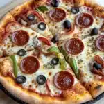 Pellet grill cast iron pizza - the image is of a large pepperoni pizza on a wooden cutting board. the pizza is round and has a golden brown crust. it is topped with slices of pepperoni, black olives, and green bell peppers. the pepperoni is arranged in a circular pattern on top of the cheese, which is melted and bubbly. there are also small pieces of sausage and other toppings scattered throughout the pizza. the cutting board is placed on a white tablecloth.