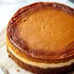 Pumpkin bread cheesecake - the image is of a round cheesecake on a wooden board. the cheesecake has a golden brown crust on top and a creamy white filling in the center. the top of the cheesecake is smooth and shiny, and it appears to be freshly baked. the board is placed on a white marble countertop, and there is a black plate in the background. the overall aesthetic of the image is elegant and sophisticated.
