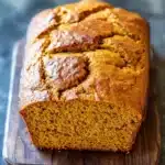 Pumpkin bread gf - the image shows a freshly baked loaf of bread on a wooden cutting board. the bread appears to be golden brown in color and has a crumbly texture. it is cut in half, revealing the soft, fluffy interior. the cutting board is placed on a dark, textured surface, possibly a table or countertop. the background is blurred, making the bread the focal point of the image.