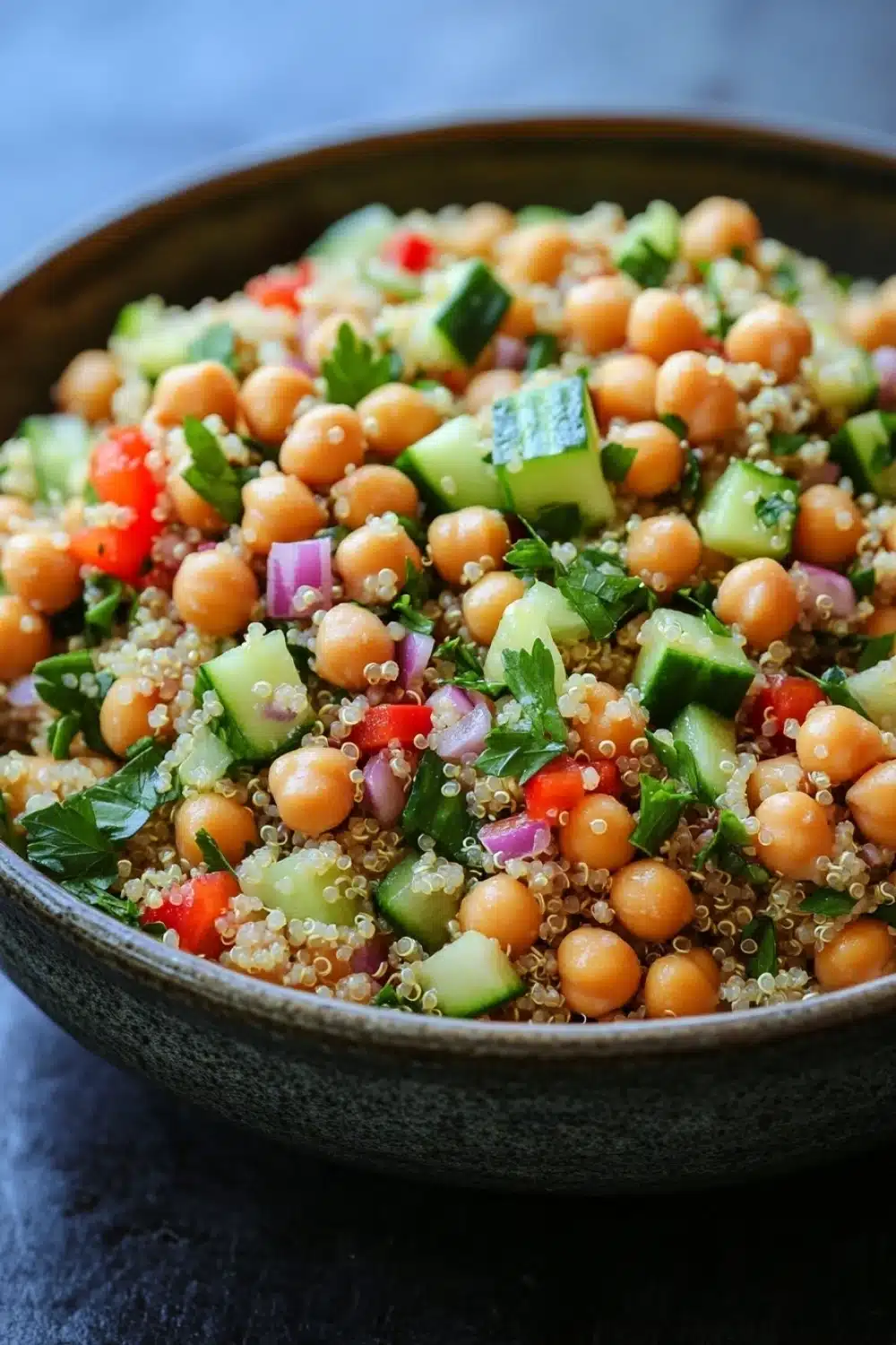 Quinoa and chickpea salad - the image is a close-up of a bowl of quinoa salad. the bowl is made of dark brown ceramic and is placed on a dark grey countertop. the salad is made up of small, round chickpeas, sliced cucumbers, red onions, and green herbs. the quinoa is a light brown color and appears to be cooked until it is soft and fluffy. the vegetables are arranged in a colorful and appetizing manner, with some overlapping each other. the overall appearance of the salad is fresh and vibrant.