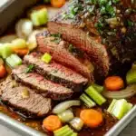 Roast beef marinade slow cooker - the image shows a rectangular baking dish with a large piece of cooked meat on it. the meat appears to be a roast beef, with a dark brown color and a shiny texture. it is garnished with chopped parsley and sliced onions. the dish is served on a bed of sliced carrots and celery, and there is a drizzle of sauce on top. the background is a white countertop.