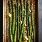 Roasted asparagus garlic - the image shows a baking tray with cooked asparagus spears on it. the tray is made of metal and has a rustic, weathered appearance. the spears are arranged in a single layer on the tray, with some of them overlapping each other. there are also small pieces of lemon wedges scattered around the tray. the background is a dark wooden table, and the tray is lined with parchment paper. the overall color scheme of the image is warm and earthy.