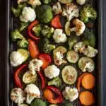 Roasted vegetables in ninja air fryer - the image shows a tray of roasted vegetables on a wooden table. the tray is rectangular in shape and has a dark brown color. the vegetables are arranged in a single layer on the tray, with some overlapping each other. on top of the vegetables, there are roasted cauliflower, broccoli, zucchini, and red bell peppers. there are also slices of orange and cucumber scattered throughout the tray. the background is blurred, but it appears to be a kitchen countertop.