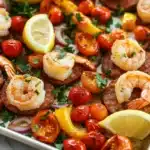 Sausage and shrimp sheet pan meal - the image shows a tray of cooked shrimp and sausage. the shrimp are cooked to a golden brown color and are arranged in a circular pattern on top of the sausage. there are also small pieces of cherry tomatoes, red onions, and yellow bell peppers scattered throughout the dish. the dish is garnished with fresh herbs and lemon wedges. the tray is sitting on a white marble countertop.