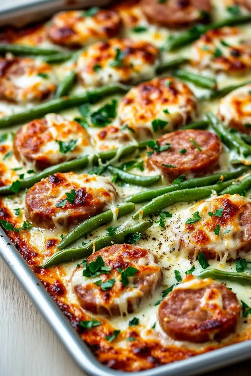 Sausage green beans alfredo sheet pan - the image shows a baking tray filled with a dish of green beans and sausage. the dish appears to be freshly baked and has a golden brown color. the green beans are long and slender, and they are arranged in a circular pattern on top of the sausage. there are also small pieces of sausage scattered throughout the dish. the sausage is golden brown and looks crispy on the outside. the sauce is melted and bubbly, and there are a few sprigs of parsley scattered on top. the tray is sitting on a wooden table, and the background is blurred.