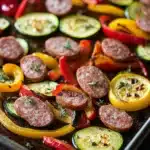 Sausage peppers and zucchini sheet pan - the image shows a baking tray filled with various types of vegetables. on the left side of the tray, there are sliced red and yellow bell peppers, zucchini, and red bell peppers. the vegetables are arranged in a circular pattern, with some overlapping each other. the tray is lined with parchment paper, and the background is dark, making the colors of the vegetables stand out. the overall appearance of the dish is colorful and appetizing.