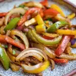 Sauteed fajita vegetables - the image is a close-up of a plate of food. the plate is round and has a blue and white speckled pattern around the edges. the food appears to be a stir-fry dish with sliced vegetables, including red, yellow, and green bell peppers. the vegetables are arranged in a circular pattern on the plate, with some overlapping each other. the colors of the vegetables are vibrant and the dish looks fresh and appetizing. the background is blurred, making the plate the focal point of the image.