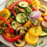 Sauteed vegetables in oven - the image is a close-up of a colorful salad on a white marble countertop. the salad is made up of various types of vegetables, including sliced zucchini, red bell peppers, yellow squash, and mushrooms. the vegetables are arranged in a circular pattern on a beige plate. there are also some sprigs of parsley scattered throughout the salad. the colors of the vegetables are vibrant and the dish looks fresh and appetizing.