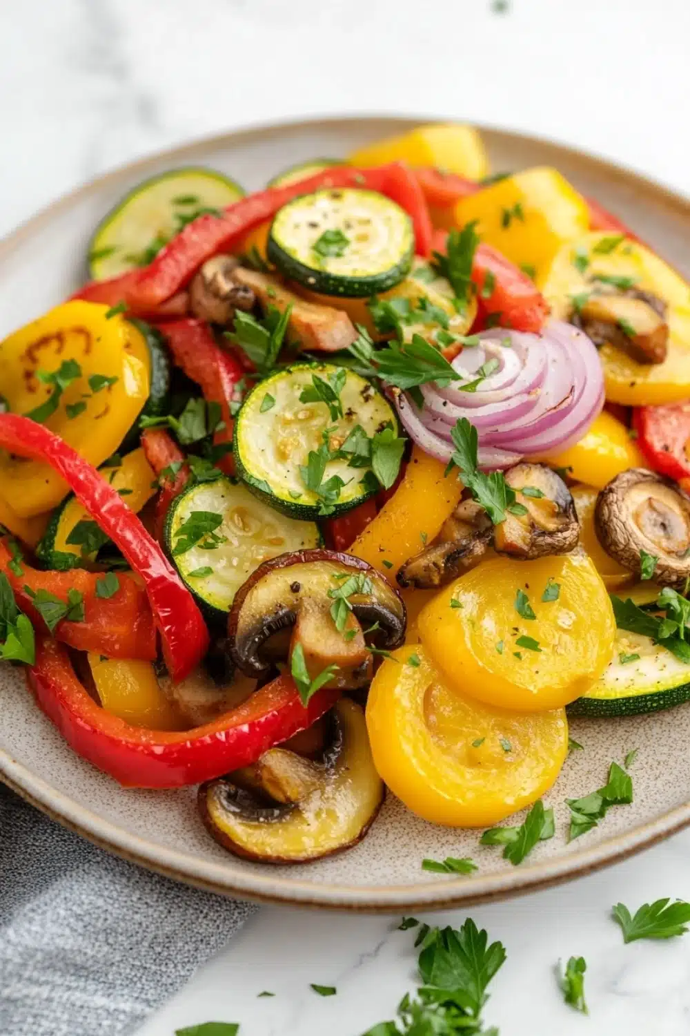 Sauteed vegetables in oven - the image is a close-up of a colorful salad on a white marble countertop. the salad is made up of various types of vegetables, including sliced zucchini, red bell peppers, yellow squash, and mushrooms. the vegetables are arranged in a circular pattern on a beige plate. there are also some sprigs of parsley scattered throughout the salad. the colors of the vegetables are vibrant and the dish looks fresh and appetizing.