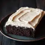 Sheet cake with cream cheese frosting - the image is a close-up of a slice of chocolate cake on a black plate. the cake appears to be moist and has a rich, dark chocolate color. the top of the cake is covered in a generous layer of white frosting, which is spread evenly across the cake. the frosting has a smooth texture and looks like it has been freshly baked. the plate is sitting on a dark wooden table, and the background is blurred, making the cake the focal point of the image.