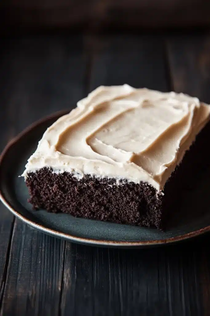 Sheet cake with cream cheese frosting - the image is a close-up of a slice of chocolate cake on a black plate. the cake appears to be moist and has a rich, dark chocolate color. the top of the cake is covered in a generous layer of white frosting, which is spread evenly across the cake. the frosting has a smooth texture and looks like it has been freshly baked. the plate is sitting on a dark wooden table, and the background is blurred, making the cake the focal point of the image.