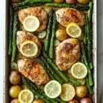 Sheet pan dinner with asparagus - the image shows a rectangular baking tray filled with cooked chicken, asparagus, potatoes, and lemons. the chicken is golden brown and appears to be seasoned with herbs and spices. the potatoes are small and round, and the lemons are bright yellow. the tray is placed on a white marble countertop, and there is a silver spoon on the right side of the image.