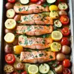 Sheet pan salmon meal - the image shows a rectangular baking tray with four pieces of salmon on it. the salmon is cooked to a golden brown color and is garnished with fresh herbs and lemon wedges. the tray is filled with roasted vegetables, including cherry tomatoes, zucchini, and potatoes. the vegetables are arranged in a circular pattern on the tray, with some overlapping each other. the background is a white marble countertop.