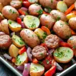 Sheet pan sausage and potatoes dinner - the image shows a baking tray filled with roasted potatoes and sausage. the potatoes are golden brown and appear to be seasoned with herbs and spices. the sausage is cut into small cubes and is arranged in a circular pattern on the tray. there are also diced red and yellow bell peppers, red onions, and yellow squash scattered throughout the dish. the tray is lined with parchment paper and is sitting on a black countertop. the overall appearance of the dish is colorful and appetizing.