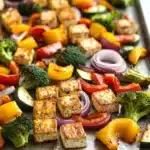 Sheet pan tofu and vegetables - the image shows a tray of grilled vegetables on a baking sheet. the vegetables are arranged in a circular pattern on the tray, with pieces of tofu, red onions, yellow bell peppers, broccoli, and zucchini scattered throughout. the tofu appears to be seasoned with herbs and spices, giving it a golden brown color. the tray is lined with parchment paper, and the vegetables are glistening with oil. the background is a white countertop.