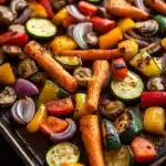 Sheet pan vegetables with carrots - the image shows a tray of roasted vegetables on a wooden table. the vegetables are arranged in a circular pattern on the tray, with some overlapping each other. the carrots are orange and appear to be seasoned with herbs and spices. there are also small pieces of red and yellow bell peppers, zucchini, and mushrooms scattered throughout the vegetables. the tray is lined with parchment paper, and the vegetables are glistening with oil. the background is dark, making the colors of the vegetables stand out.