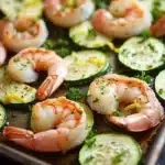 Shrimp and zucchini sheet pan dinner - the image shows a baking tray filled with cooked shrimp and zucchini. the shrimp are pink and appear to be seasoned with herbs and spices. the zucchinis are sliced and arranged in a circular pattern on the tray. the tray is lined with parchment paper and there are a few sprigs of parsley scattered around the tray, adding a pop of green color to the dish. the background is dark, making the colors of the dish stand out.