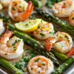Shrimp sheet pan with asparagus - the image shows a tray of cooked shrimp and asparagus on a wooden table. the shrimp are arranged in a single layer on the tray, with some of them overlapping each other. the asparaglips are bright green and appear to be seasoned with herbs and lemon wedges. the tray is lined with parchment paper, and there are a few sprigs of parsley scattered around the tray. the background is blurred, but it appears to be a kitchen countertop.