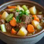 Slow cooker beef and vegetables stew - the image is a close-up of a bowl of beef stew. the bowl is made of ceramic and is sitting on a dark wooden table. the stew is a rich brown color and appears to be made with chunks of beef, carrots, potatoes, celery, and other vegetables. there is a sprig of parsley on top of the stew, adding a pop of green color to the dish. the broth is thick and thick, and the vegetables are cooked to perfection. the overall appearance is hearty and hearty.