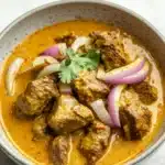 Slow cooker beef korma curry - the image is a close-up of a bowl of indian curry. the curry is a rich orange color and appears to be made with chunks of meat and vegetables. the meat is cooked in a tomato-based sauce and is garnished with sliced red onions and a sprig of cilantro. the bowl is white with a speckled pattern and is sitting on a white marble countertop.