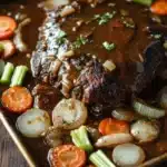 Slow cooker beef roast and gravy - the image shows a rectangular baking tray filled with a dish of beef stew. the beef is cooked medium-rare and is covered in a rich, dark brown sauce. the sauce is drizzled over the top of the beef and is garnished with chopped parsley. there are also sliced carrots, celery, and onions scattered throughout the dish. the tray is sitting on a wooden table with a white cloth napkin in the background.