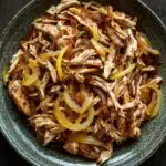 Slow cooker chicken carnitas - the image is a close-up of a plate of food. the plate is made of a dark green ceramic bowl with a textured surface. the food appears to be a dish made with shredded chicken, sliced onions, and yellow bell peppers. the chicken is cooked until tender and the onions are sliced into thin strips. the bell peppers are bright yellow and appear to be seasoned with herbs and spices. the dish is garnished with a sprinkle of sesame seeds. the background is a dark wooden table.