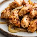 Slow cooker chicken tenderloin - the image shows a plate of grilled chicken wings on a white marble countertop. the wings are golden brown and appear to be seasoned with herbs and spices. they are arranged in a circular pattern on the plate, with some of the wings overlapping each other. there are also some sliced onions scattered around the plate. the plate is round and has a dark brown rim. the background is blurred, making the plate the focal point of the image.