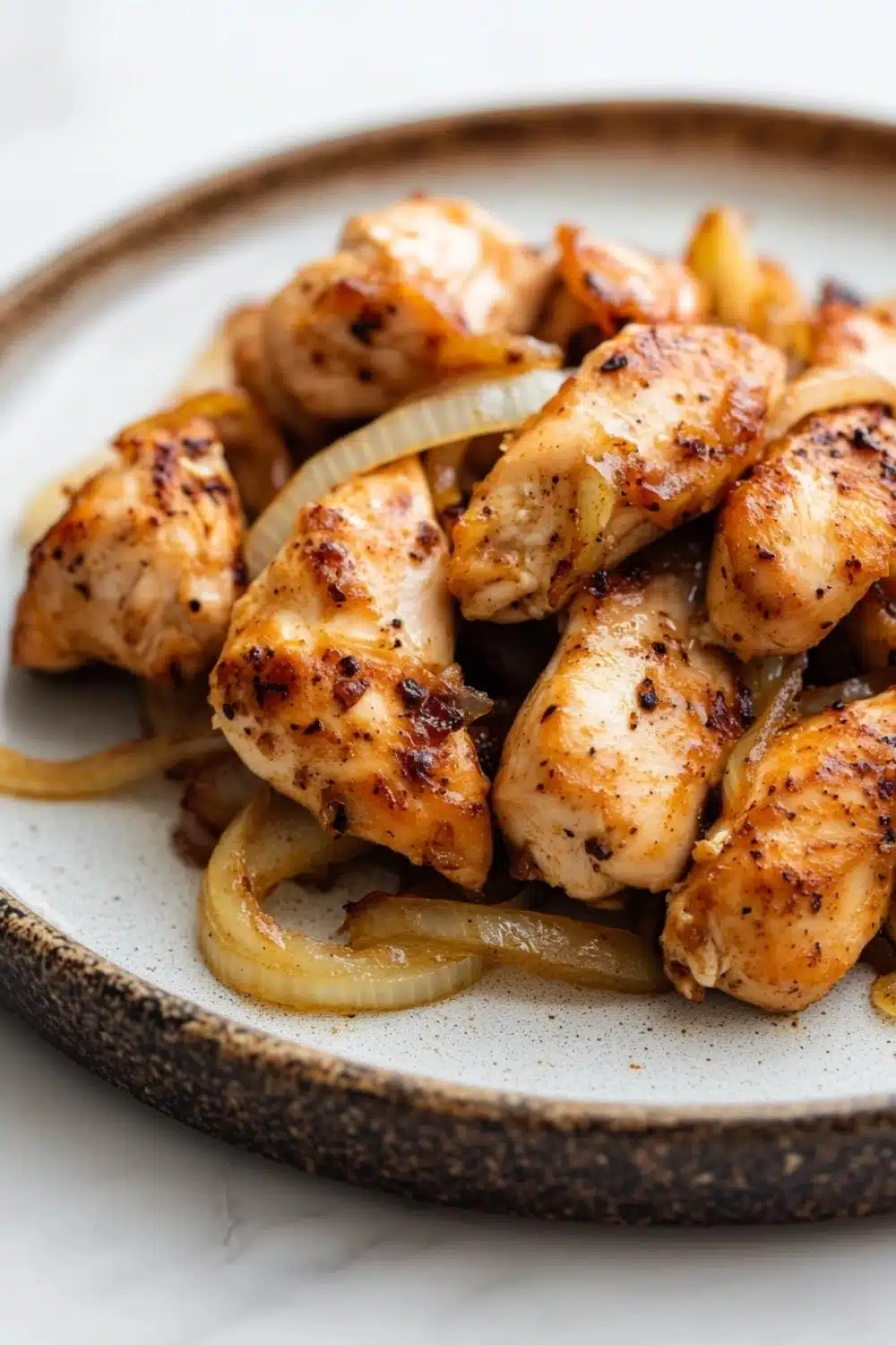 Slow cooker chicken tenderloin - the image shows a plate of grilled chicken wings on a white marble countertop. the wings are golden brown and appear to be seasoned with herbs and spices. they are arranged in a circular pattern on the plate, with some of the wings overlapping each other. there are also some sliced onions scattered around the plate. the plate is round and has a dark brown rim. the background is blurred, making the plate the focal point of the image.