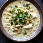 Slow cooker white chicken chili heavy cream - the image is a close-up of a bowl of soup. the soup appears to be a creamy white color with chunks of chicken and beans mixed in. the bowl is made of ceramic and has a brown rim. there are also some fresh cilantro leaves scattered on top of the soup, adding a pop of green color to the dish. the background is a dark grey countertop.