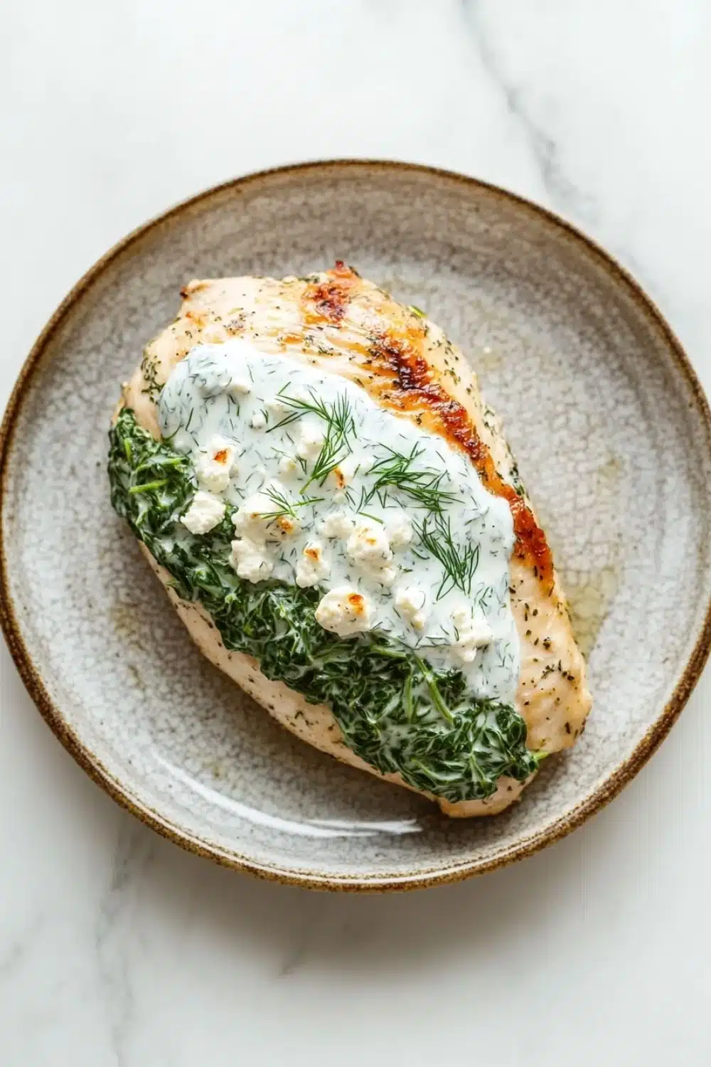 Spinach stuffed chicken breast greek yogurt - the image shows a plate of food on a white marble countertop. the plate is round and has a light brown color with a speckled pattern. on top of the plate, there is a piece of grilled chicken breast covered in a creamy white sauce. the chicken breast is garnished with fresh dill leaves and crumbled feta cheese. the dish appears to be freshly cooked and ready to eat.
